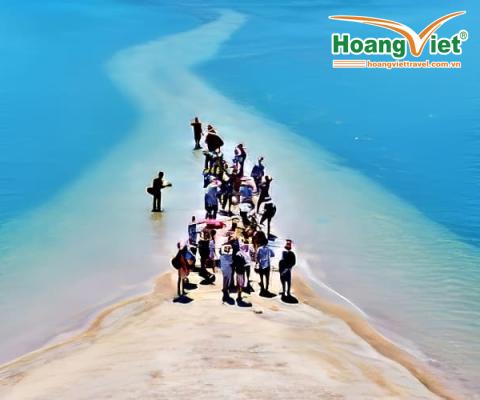 Một nhóm người tụ tập trên một dải cát hẹp bao quanh bởi làn nước xanh thẳm dưới bầu trời trong xanh. Khung cảnh vui tươi này truyền tải cảm giác phiêu lưu và khám phá.