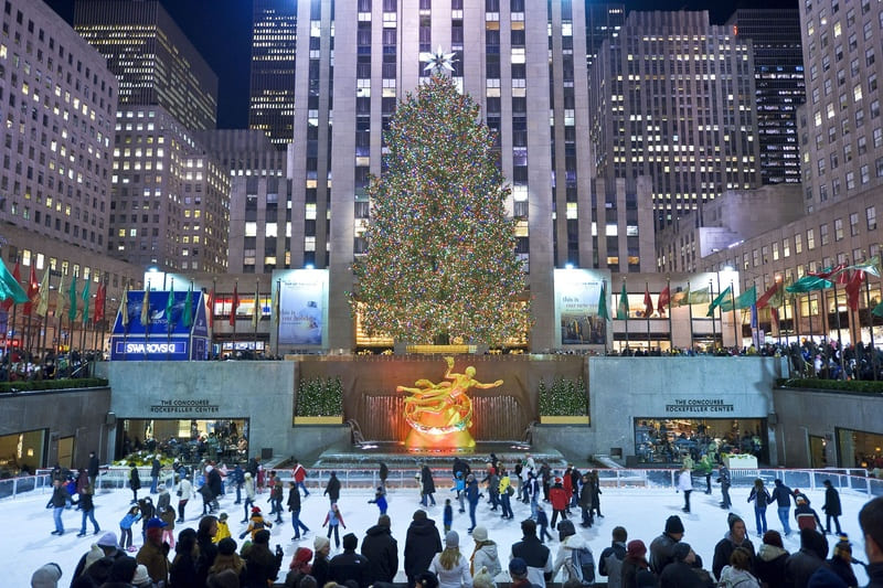 Một sân trượt băng nhộn nhịp tại Rockefeller Center với những người trượt băng đang di chuyển. Một cây thông Noel khổng lồ, được thắp sáng rực rỡ, sừng sững giữa những ánh đèn rực rỡ của thành phố.