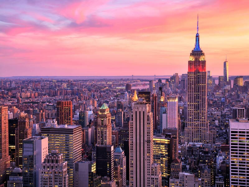 Cảnh quan thành phố New York lúc hoàng hôn, nổi bật với tòa nhà Empire State Building dưới bầu trời rực rỡ sắc hồng và cam. Khung cảnh thành phố toát lên vẻ thanh bình và hùng vĩ.