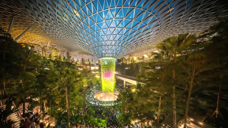 Thác nước trong nhà sống động tại sân bay Jewel Changi của Singapore, được bao quanh bởi cây xanh tươi tốt dưới mái kính lớn dạng lưới, tạo nên một không gian thanh bình và đầy cảm hứng.