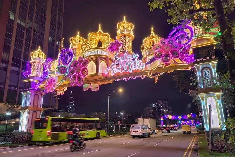 Cổng vòm ánh sáng rực rỡ với các họa tiết hoa lá và hình học mang tên Hari Raya Aidilfitri trải dài trên một con phố nhộn nhịp vào ban đêm, tạo nên một bầu không khí lễ hội sống động.
