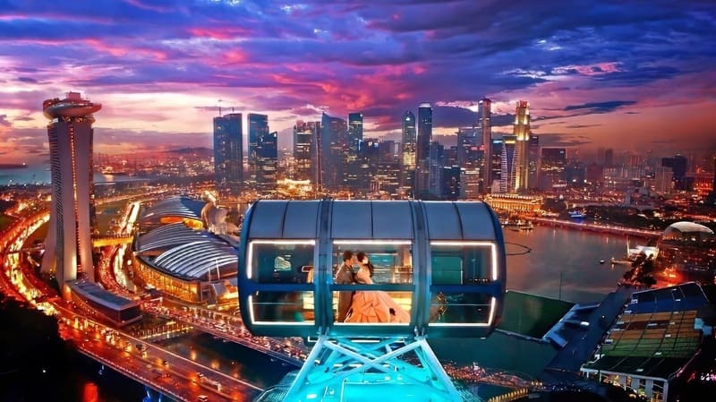 Một cặp đôi ôm nhau bên trong một cabin kính trên vòng đu quay, với khung cảnh đường chân trời rực rỡ ánh đèn của Singapore và bầu trời hoàng hôn rực rỡ phía sau, truyền tải một thông điệp lãng mạn.