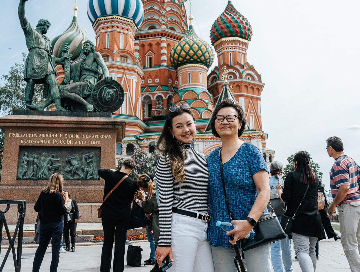 Hai người phụ nữ mỉm cười trước Nhà thờ Thánh Basil, Moscow. Những mái vòm đầy màu sắc và bức tượng đồng nổi bật giữa khu vực du lịch nhộn nhịp.