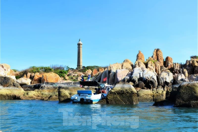 Kỳ thú những cung đường bộ xuyên biển - Ảnh 1. Một chiếc thuyền neo đậu gần bờ đá, phía sau là ngọn hải đăng cao vút dưới bầu trời xanh trong vắt.