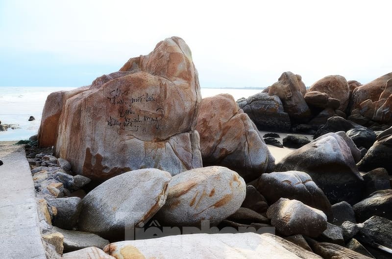 Kỳ thú những cung đường bộ xuyên biển - Ảnh 3. Những tảng đá lớn ven biển có khắc chữ gần đại dương, được bao quanh bởi những viên đá nhỏ hơn dưới bầu trời trong xanh, tạo nên một khung cảnh thanh bình.