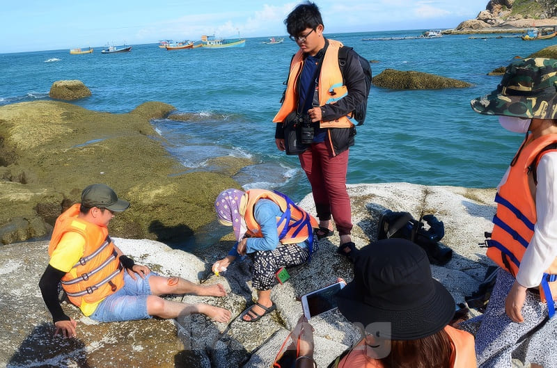 Kỳ thú những cung đường bộ xuyên biển - Ảnh 4. Du khách mặc áo phao cứu sinh giúp đỡ một người đàn ông bị thương trên bờ đá ven biển, phía sau là những chiếc thuyền dưới bầu trời xanh trong vắt.