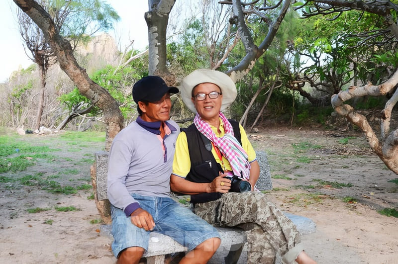 Kỳ thú những cung đường bộ xuyên biển - Ảnh 6. Hai người đàn ông ngồi trên ghế đá công viên, một người cầm máy ảnh. Xung quanh họ là cây cối và thảm thực vật xanh tươi, tận hưởng một khoảnh khắc yên bình.