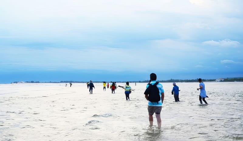 Kỳ thú những cung đường bộ xuyên biển - Ảnh 8. Mọi người đi bộ qua vùng nước nông trên một bãi biển rộng lớn dưới bầu trời nhiều mây, tạo nên một khung cảnh thanh bình và đầy phiêu lưu.
