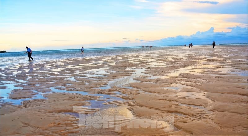 Kỳ thú những cung đường bộ xuyên biển - Ảnh 9. Mọi người tản bộ trên một bãi biển rộng lớn, phản chiếu ánh sáng lúc hoàng hôn, với bầu trời đầy màu sắc và đường chân trời xa xăm, tạo nên một khung cảnh thanh bình, tĩnh lặng.