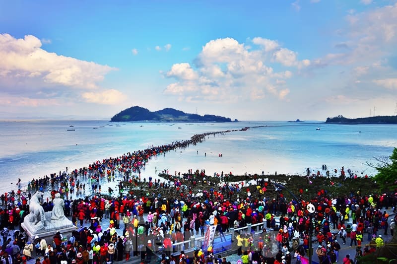 Kỳ thú những cung đường bộ xuyên biển - Ảnh 14. Crowds gather on a narrow path revealed by low tide, connecting to an island under a clear blue sky with scattered clouds.