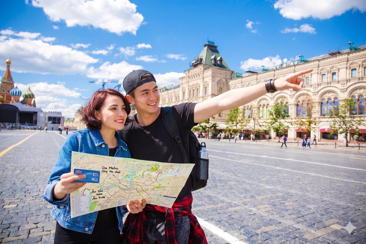 Một cặp đôi tươi cười, tay cầm bản đồ, đứng trong một quảng trường rực rỡ ánh nắng với những tòa nhà cổ kính. Người đàn ông chỉ tay về phía trước, như muốn chỉ hướng hoặc gợi lên điều gì đó thú vị.
