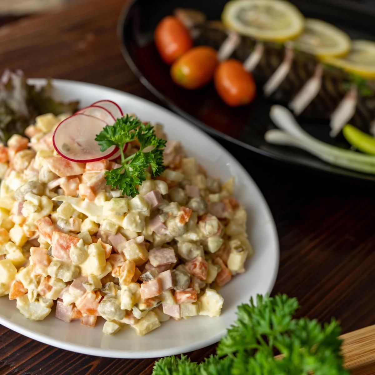 Một đĩa salad tươi ngon trên đĩa trắng với rau củ thái hạt lựu, trang trí bằng rau mùi tây và lát củ cải. Phía sau là cá nướng chanh và cà chua.