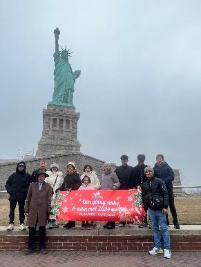 Một nhóm người tạo dáng chụp ảnh với một biểu ngữ màu đỏ rực rỡ trước Tượng Nữ thần Tự do vào một ngày nhiều mây, truyền tải một tâm trạng vui vẻ, hân hoan.