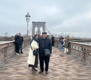 Một cặp đôi tươi cười đứng trên lối đi bằng gỗ của cầu Brooklyn vào một ngày nhiều mây. Những người đi bộ khác có thể được nhìn thấy ở phía sau, đang tận hưởng khung cảnh.