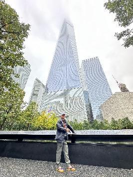 Một người đứng trước một tòa nhà chọc trời cao vút, phản chiếu ánh sáng, bao quanh là những tòa nhà nhỏ hơn và cây cối. Bầu trời nhiều mây, tạo nên một bầu không khí thanh bình.