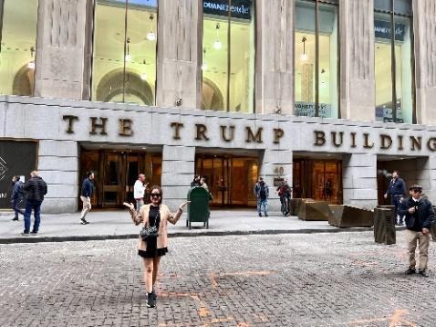 Một người phụ nữ đứng mỉm cười trên con phố lát đá cuội trước tòa nhà Trump. Mọi người đi lại gần đó, tạo nên một bầu không khí sống động và nhộn nhịp.