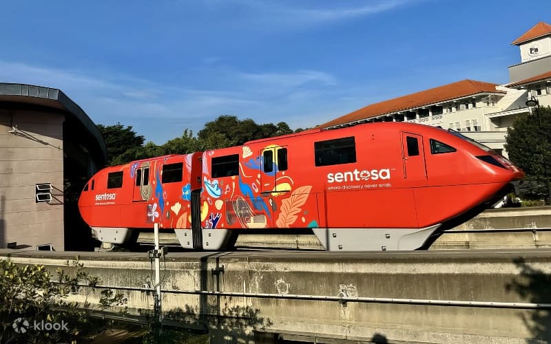 Một đoàn tàu điện một ray Sentosa màu đỏ rực rỡ, được trang trí bằng những tác phẩm nghệ thuật đầy màu sắc, đang di chuyển trên đường ray trên cao dưới bầu trời xanh trong vắt. Cây xanh tươi tốt và các tòa nhà hiện lên ở phía xa.